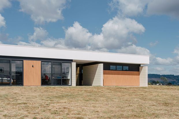 Single-storey contemporary house viewed from the front across an open grassy site. The building has a long, low flat roof, extensive dark-framed glazing, pale rendered or masonry walls, and warm vertical timber-clad feature panels around the entry and garage-like frontage. The composition is highly horizontal and minimalist, with a covered recessed entry and a broad projecting roof plane. Blue sky and distant landscape form the background.
