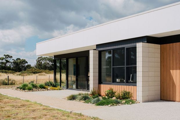 Exterior view of a low, single-storey contemporary residence with a broad flat roof, clean rectilinear lines, white upper facade panels, pale masonry cladding, dark-framed glazing, and warm vertical timber wall cladding beside the entry. A sheltered entry porch opens onto a concrete path, with restrained native-style landscaping and an open rural-looking setting.