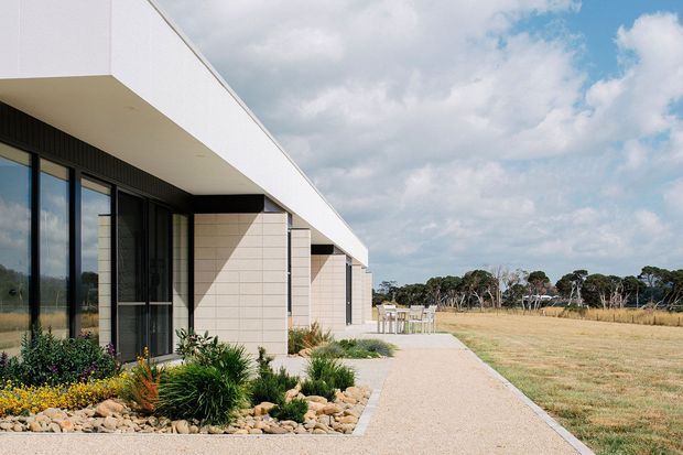 Exterior view of a long, low-slung contemporary building with crisp white walls, dark full-height glazing, projecting flat roof planes, and pale rendered or masonry feature bays. A landscaped rock garden borders the facade, while a broad gravel or concrete-paved walkway leads toward an open grassy site and outdoor seating area.