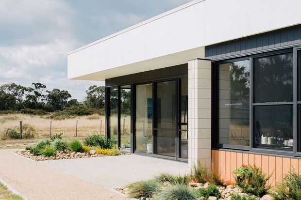 Contemporary single-storey building exterior with a clean flat-roofed form, broad white rendered or panelled fascia, dark-framed floor-to-ceiling glazing, pale masonry cladding, and warm vertical timber-look lower wall cladding. The entrance is set beneath a deep projecting canopy, opening onto a paved path and low, drought-tolerant landscaped garden. A rural field and trees are visible beyond the property.