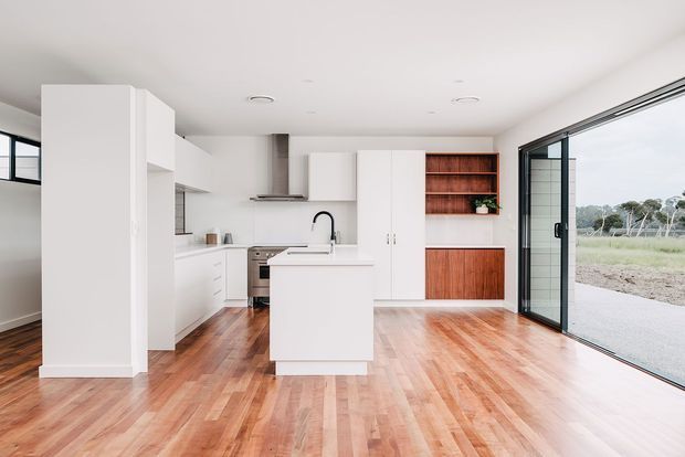 Bright open-plan kitchen with white flat-panel cabinetry, a central island, stainless-steel appliances, and open timber shelving with matching timber cabinetry. Warm timber flooring extends through the room, while large black-framed sliding glass doors provide views to the outdoors.