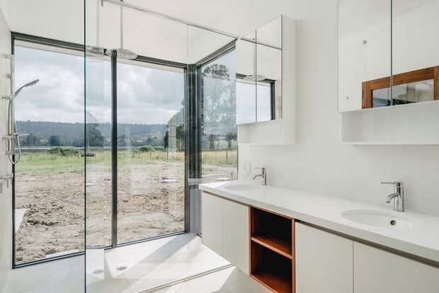 Bright contemporary bathroom with a large frameless glass walk-in shower, double vanity, white benchtop and cabinetry, and a timber-lined open shelf. Floor-to-ceiling glazing provides views toward a rural landscape, while mirrored cabinets increase the sense of space.