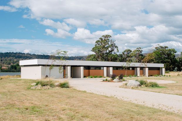 A low, single-storey contemporary residence with a strongly horizontal composition and broad flat roof. The long facade combines pale rendered or masonry volumes with dark-framed openings, warm timber-toned wall panels, and a sheltered central entry or courtyard zone. Set within a landscaped rural or semi-rural site, the house is approached by a pale gravel driveway and surrounded by native grasses, scattered rocks, and mature trees. The image has bright natural daylight and an open, calm atmosphere.