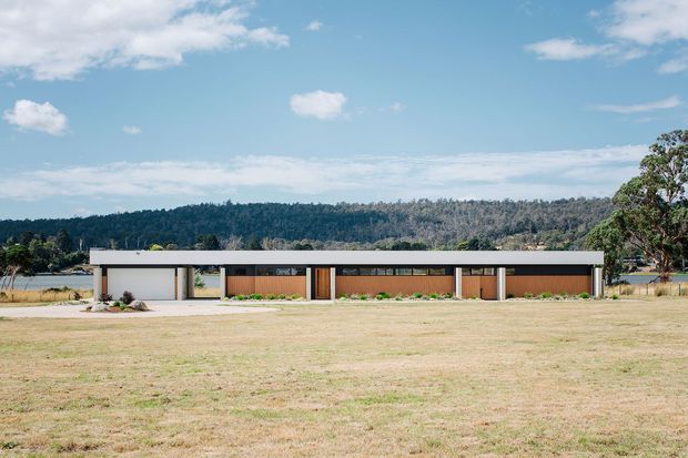 A low, elongated single-storey contemporary residence viewed from across a broad dry lawn. The building has a strong flat roofline, white rendered or painted walls, dark timber-toned recessed facade sections, extensive glazing, and a simple landscaped frontage. A wooded hillside and open landscape form the background.