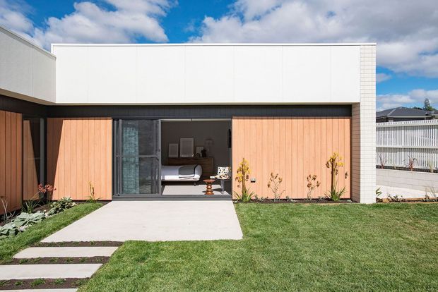 A low, minimalist contemporary house facade with a crisp white upper volume, warm vertical timber cladding around a recessed entry, and dark-framed glazing. A concrete path crosses a neatly maintained lawn and simple planted garden beds; the open doorway reveals part of a bedroom interior. The image is photographed in bright daylight against a partly cloudy sky.