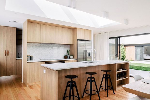 Bright contemporary kitchen with extensive light timber cabinetry, a large stone-topped island with seating, integrated stainless-steel appliances, and a marble-look splashback. Skylights and large glazed doors create an airy connection to the landscaped outdoor area, while a dining space is visible at right.