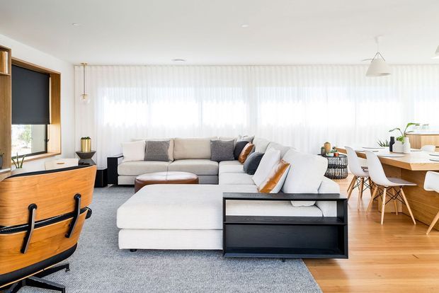 Bright open-plan living and dining area with a large light-grey sectional sofa, timber flooring, sheer full-width curtains, and contemporary pendant lighting. A timber media console and built-in shelving sit to the left, while a timber dining table with white chairs is visible to the right. The room has a clean, airy contemporary feel with warm wood accents and abundant natural light.