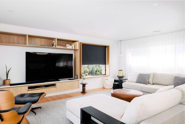 Bright contemporary living room with a large cream sectional sofa, black rectangular coffee table, and cognac leather ottoman. A timber built-in media wall provides open shelving, cabinetry, and a wall-mounted television. Timber flooring, a large pale rug, sheer floor-to-ceiling curtains, and a window with a dark roller blind complete the clean, warm space. A black leather lounge chair with a timber shell sits in the foreground.