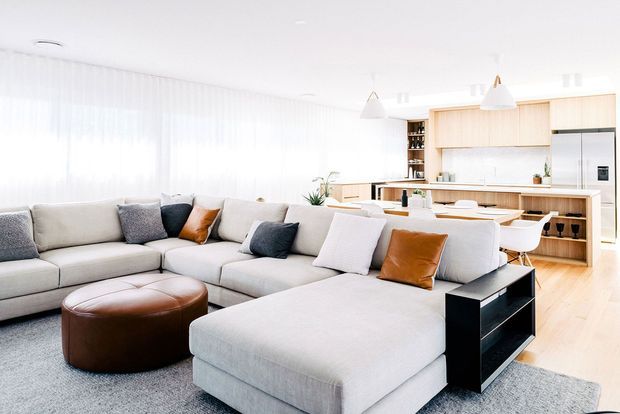 Bright open-plan living and kitchen interior with a large light-grey sectional sofa, chaise lounge, leather ottoman, and black side shelving. The adjoining kitchen has pale timber cabinetry, a long island with seating, integrated appliances, pendant lights, and light timber flooring. Sheer curtains diffuse abundant natural light across the contemporary space.