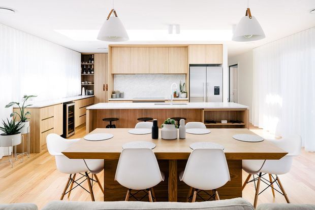 Bright open-plan kitchen and dining area with pale timber cabinetry, a large central island, integrated stainless-steel appliances, and a timber dining table surrounded by white contemporary chairs. Sheer curtains, pendant lights, light timber flooring, and indoor plants create an airy, warm, refined interior.