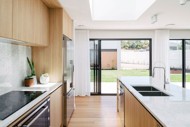 Bright contemporary kitchen with a long central island, pale timber cabinetry, white marble-look benchtops and splashback, integrated stainless-steel appliances, and dark-framed floor-to-ceiling glazing opening to a grassed backyard. Sheer curtains, skylight-like ceiling recesses, and abundant natural light create an airy, refined interior.