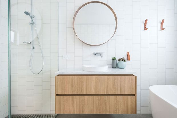 Bright contemporary bathroom featuring a floating timber vanity with an integrated white basin, white rectangular wall tiles, a frameless glass shower, round timber-framed mirror, wall-mounted tapware, and a partially visible freestanding bathtub. Small leather-toned wall hooks and potted plants add subtle warmth.