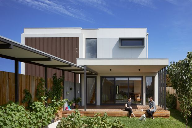 A contemporary two-storey residential house viewed from the landscaped rear garden. The white rendered facade has clean rectilinear volumes, a projecting flat-roofed ground-floor terrace, dark-framed glazing, and a tall vertical timber-clad feature wall. A covered outdoor deck with slender steel posts sits beside garden planting and timber fencing.