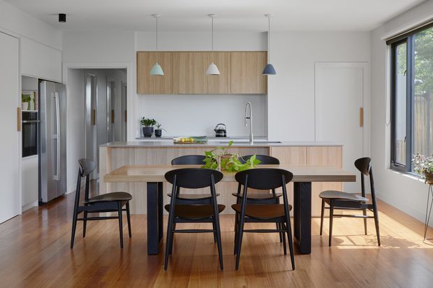 Bright open-plan kitchen and dining area with a central island, integrated white cabinetry, light timber overhead joinery, stainless-steel appliances, and a long dark dining table surrounded by black chairs. Pendant lights hang above the island and table, while large windows bring in abundant natural light. Warm timber flooring and restrained contemporary detailing create a clean, inviting interior.