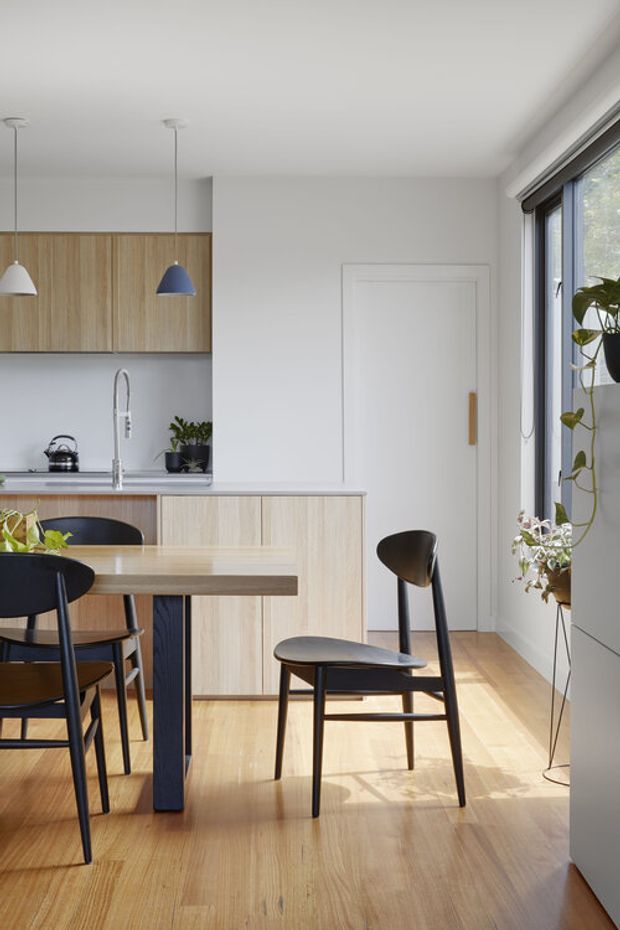 A bright, compact open-plan kitchen and dining area with pale timber cabinetry, a light timber dining table and floor, and black dining chairs. The kitchen has a simple grey backsplash, integrated cabinetry, a stainless tap and sink, and minimalist pendant lights, including a blue accent pendant. White walls, a white paneled door, and full-height glazing create an airy contemporary interior with indoor plants and strong natural light.