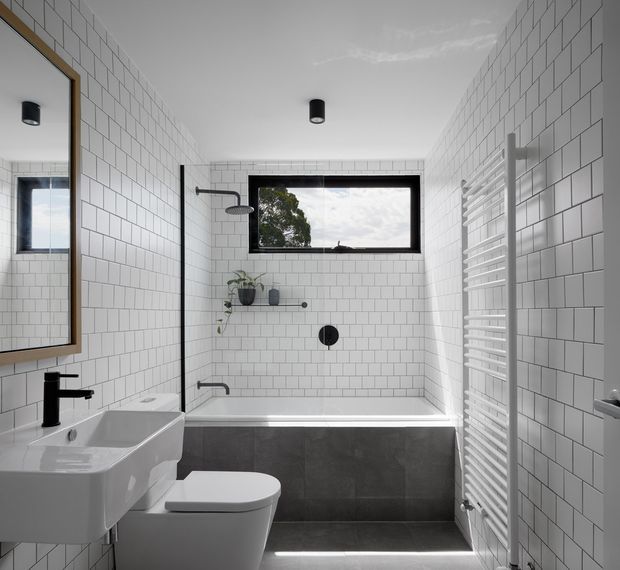 A compact contemporary bathroom with white square wall tiles, a built-in bathtub and shower, wall-mounted white basin, toilet, black tapware and fittings, a timber-framed mirror, and a white heated towel rail. A high horizontal window provides natural light, while dark grey bath-side tiles and black fixtures create contrast.