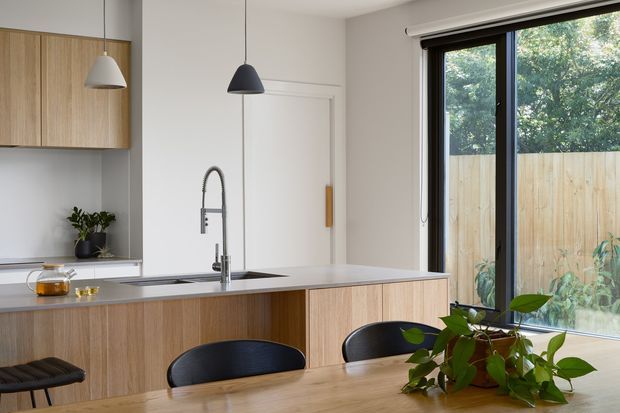 Bright, refined contemporary kitchen and dining area with flat-panel light timber cabinetry, a pale stone island benchtop, matte black pendant lights, black-framed floor-to-ceiling glazing, and a timber dining table with black chairs. Indoor plants and a timber privacy fence visible outside add a natural element.