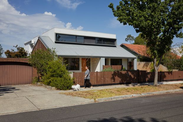 Street-level exterior view of a contemporary suburban residence with a steep metal gable roof, a raised box-like upper volume with horizontal glazing, timber-toned cladding or screening, and a separate garage. A front fence, paved driveway, footpath, mature tree, and neighbouring roof are visible.
