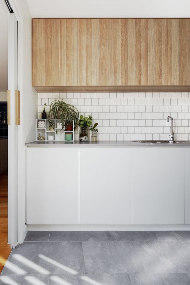 A compact contemporary kitchen with flat-panel white lower cabinetry, warm vertical-grain timber overhead cupboards, a white subway-tile splashback, and a slim grey benchtop. A chrome gooseneck tap and undermount sink sit beneath the cabinetry, while potted plants and small decorative items add greenery along the benchtop. Large grey floor tiles and strong daylight complete the clean, understated scheme.