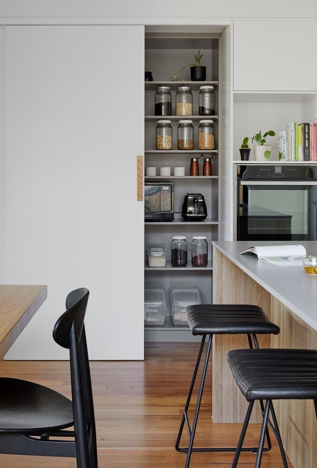 Contemporary kitchen interior featuring a tall open pantry with neatly arranged glass storage jars, integrated black oven, white flat-panel cabinetry, and a timber kitchen island with a pale benchtop. Black upholstered bar stools and dining furniture contrast with the light cabinetry, while timber flooring adds warmth. The space is orderly, functional, and refined.