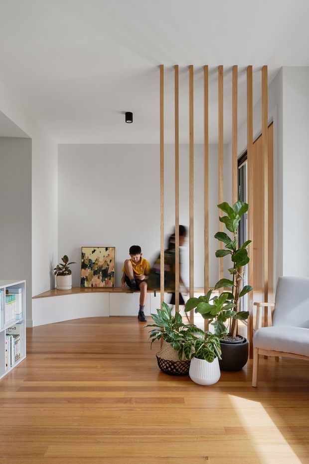 Bright contemporary living area with warm timber flooring, a built-in low timber bench and cabinetry, and a vertical timber-screen partition extending to the ceiling. Large windows bring in strong natural light, while indoor plants and a pale upholstered armchair add softness. A child is seated on the built-in bench, and a small open bookshelf is visible at the left.