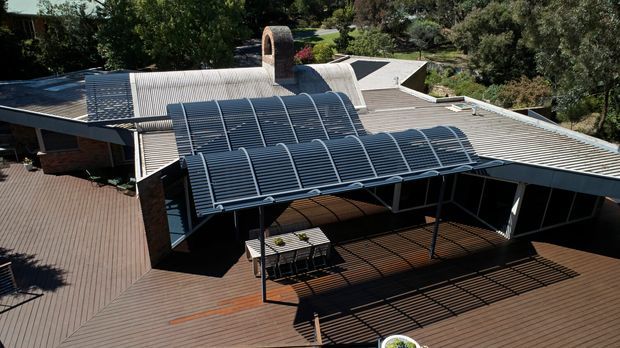 Aerial view of a low-profile modern residence with extensive corrugated metal roofing, curved translucent roof canopies, a brick chimney, and a large timber deck with a shaded outdoor dining area. The home is set within mature landscaped vegetation.