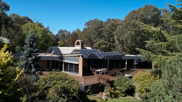 Exterior view of a low-rise residential building set within mature landscaped trees. The house has a long, low horizontal form, dark walls, extensive glazing, a broad timber-toned deck, and a distinctive curved translucent roof structure with a small brick chimney or arched feature. Clear blue sky and dense garden planting surround the dwelling.