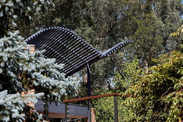 Exterior architectural detail of a contemporary pavilion or entrance canopy set within dense established vegetation. The defining feature is a dramatic curved, wing-like slatted roof structure supported by slender dark posts, with brick and dark wall elements partially visible behind it.