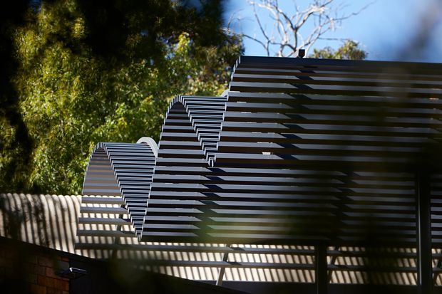 Close-up exterior view of a contemporary building facade featuring layered horizontal dark metal slats forming angular, folded roof or screen profiles. Strong sunlight creates striped shadows across the wall and structure, with mature trees visible in the background.