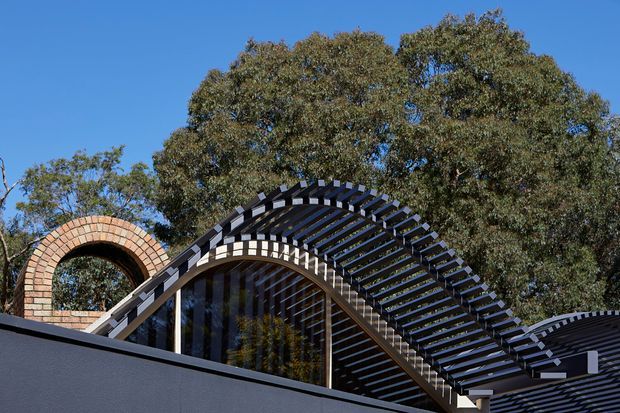 Exterior architectural detail showing a dramatic curved, wave-like roof or canopy formed by repeated dark structural ribs over a glazed facade. A circular brick arch feature is visible to the left, with mature trees and blue sky behind the building. The image emphasizes the sculptural roofline and exterior structure.