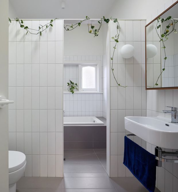 Bright, compact contemporary bathroom with white square wall tiles, grey tiled flooring, a wall-mounted white basin, toilet, bathtub, chrome fixtures, and a large timber-framed mirrored cabinet. Trailing green plants add an organic accent, while a deep blue hand towel provides contrast.