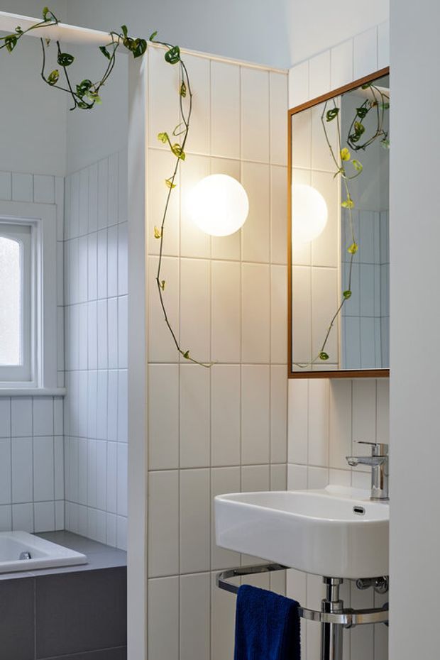 A bright, compact bathroom featuring white vertically arranged wall tiles, a freestanding wall-mounted basin with exposed chrome plumbing, a timber-framed mirrored cabinet, and a round wall light. A trailing plant adds a natural accent. A bathtub is partially visible beside a frosted window.