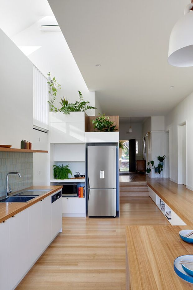 Bright open-plan kitchen viewed from the dining area, with white cabinetry, timber benchtops and extensive light timber flooring. A stainless-steel refrigerator sits within a partial-height joinery wall, while a tiled splashback, open shelving, indoor plants and a long circulation zone lead toward a raised living area and exterior doorway. Natural light enters through high windows with vertical blinds, creating an airy contemporary interior.
