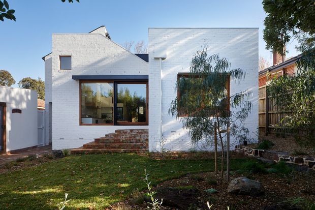 Exterior view of a compact white-painted brick residence with a contemporary flat-roofed addition beside an older pitched-roof section. The facade features large timber-framed windows, brick steps, a small front lawn, planted garden beds, and timber fencing. A neighbouring brick house is visible at the right edge.