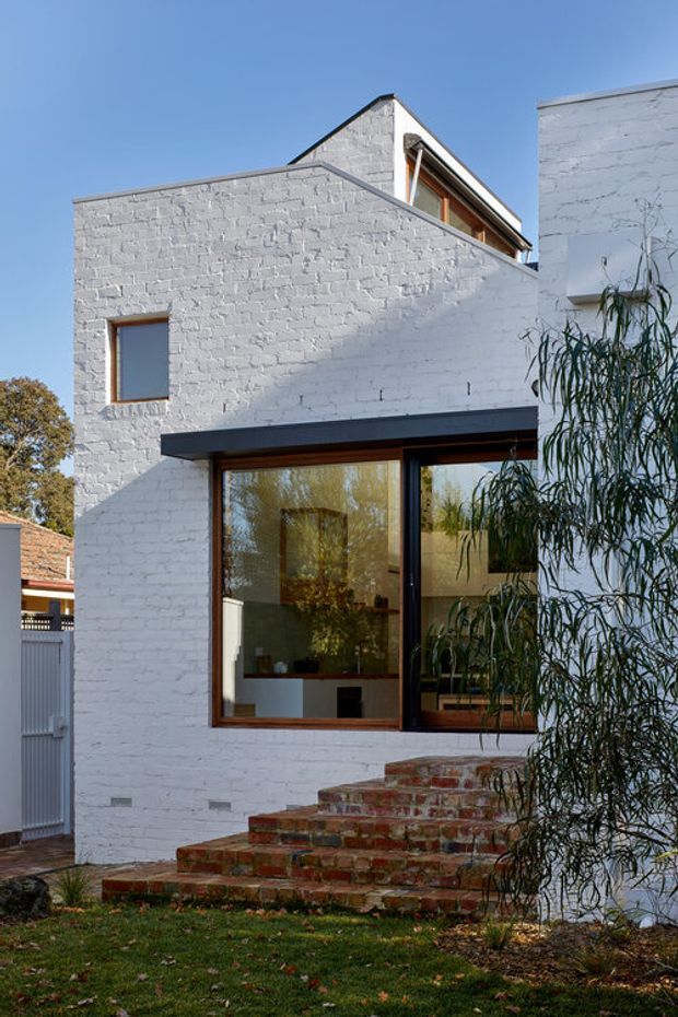 Exterior view of a compact multi-level residence with white-painted brick walls, dark metal roof and window trim, and warm timber-framed glazing. Broad brick steps lead up to a large ground-floor window, through which a minimal interior and indoor planting are visible. The house has a modern, angular composition with a pitched roof element behind a flat-roofed front volume, set in a leafy suburban garden.
