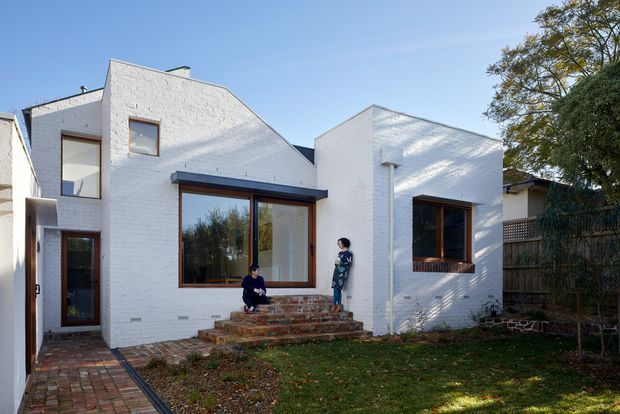 Exterior view of a low-rise white-painted brick house with a distinctive stepped and pitched roofline, dark timber-framed windows, and a recessed entry along a brick-paved walkway. Brick steps lead to a small front garden and lawn, with neighbouring buildings and mature trees visible around the property.