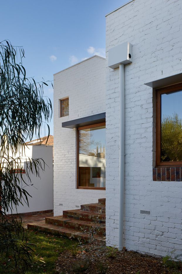 Exterior view of a compact contemporary home with white-painted brick walls, crisp rectilinear volumes, timber-framed windows, and an inset entry reached by rustic brick steps. A white downpipe and projecting horizontal window awning are visible, with landscaped garden planting in the foreground and neighbouring residential elements beyond.