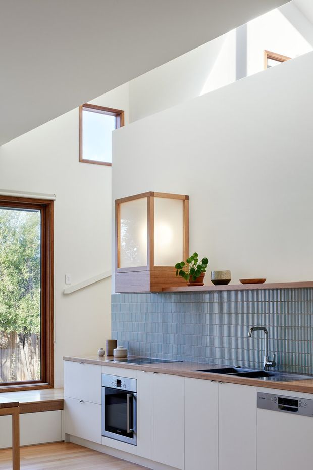 Bright, compact contemporary kitchen with a high sloping ceiling and clerestory-style windows. White flat-panel cabinetry runs beneath a muted blue vertically ribbed tile splashback, paired with a warm timber benchtop and integrated stainless-steel appliances. Timber-framed glazing, pale timber flooring, and a built-in timber display cabinet add warmth. A small sink is positioned at the right, with natural light and leafy outdoor views entering through the windows.