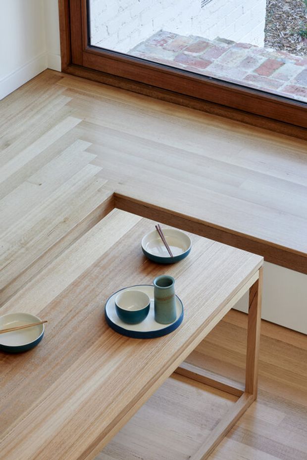 A close-up interior view of a light timber-finished seating area beside a large timber-framed window. A built-in window bench or ledge and a simple timber coffee table display ceramic bowls and cups. Pale timber flooring and joinery create a warm, minimal setting, with a brick-paved exterior visible through the glazing.
