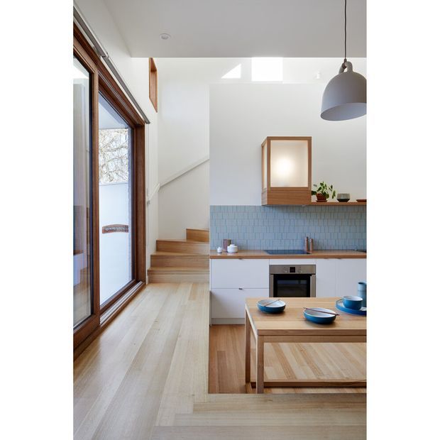 Bright open-plan kitchen and dining area with a light timber floor, white cabinetry, pale blue tiled splashback, timber-framed glazing, and a timber dining table. A compact timber staircase rises beside the kitchen, while a large grey pendant light and natural light from the glazing create an airy, contemporary atmosphere.