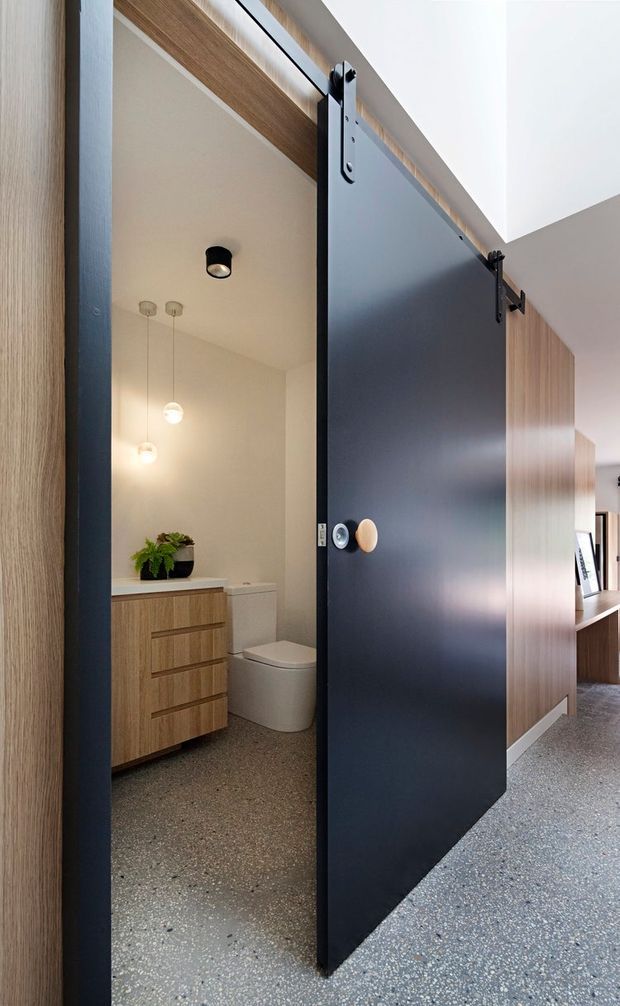 A contemporary interior view looking toward a compact bathroom through a partially open matte dark sliding barn-style door. The bathroom has a warm timber vanity with drawers, white basin area, white toilet, terrazzo-like speckled flooring, white walls, and pendant lighting. The surrounding hall or living area features matching timber wall panelling and a built-in display niche.