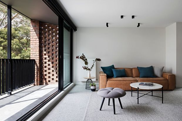 A contemporary living room with a tan leather sofa, dark teal cushions, a round white coffee table, and a sculptural grey ottoman on a pale carpet. Full-height black-framed glazing opens to a narrow balcony or terrace, while a perforated red brick screen and black balustrade provide architectural detail. The room is bright and minimally furnished, with track lighting and a potted plant.