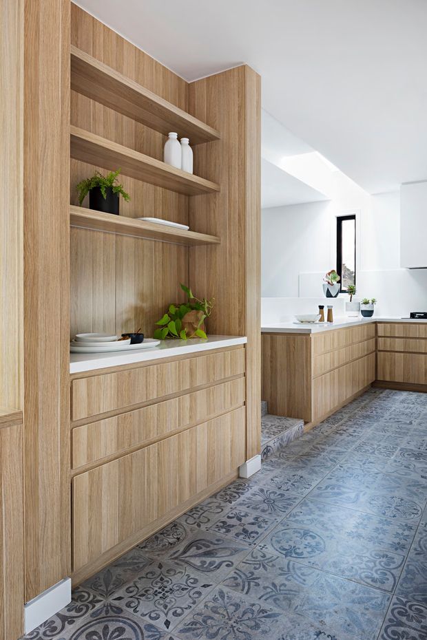 Bright contemporary kitchen with extensive light timber cabinetry and open display shelving. The foreground features a tall built-in shelving and drawer unit with white benchtop, while the rear has an L-shaped kitchen with matching timber lower cabinetry, white surfaces, a narrow vertical window, and a skylight. Decorative patterned blue-grey floor tiles add visual contrast.