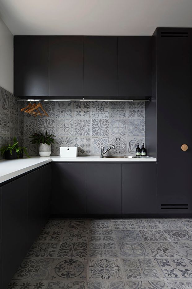 Compact L-shaped kitchen with matte charcoal-black flat-panel cabinetry, a white benchtop, and a decorative grey patterned tile splashback continuing onto the floor. The space includes a stainless-steel sink and tap, integrated dark appliance cabinetry, open hanging rail storage, and a few potted plants. The overall look is dark, graphic, and contemporary with an eclectic patterned-tile feature.