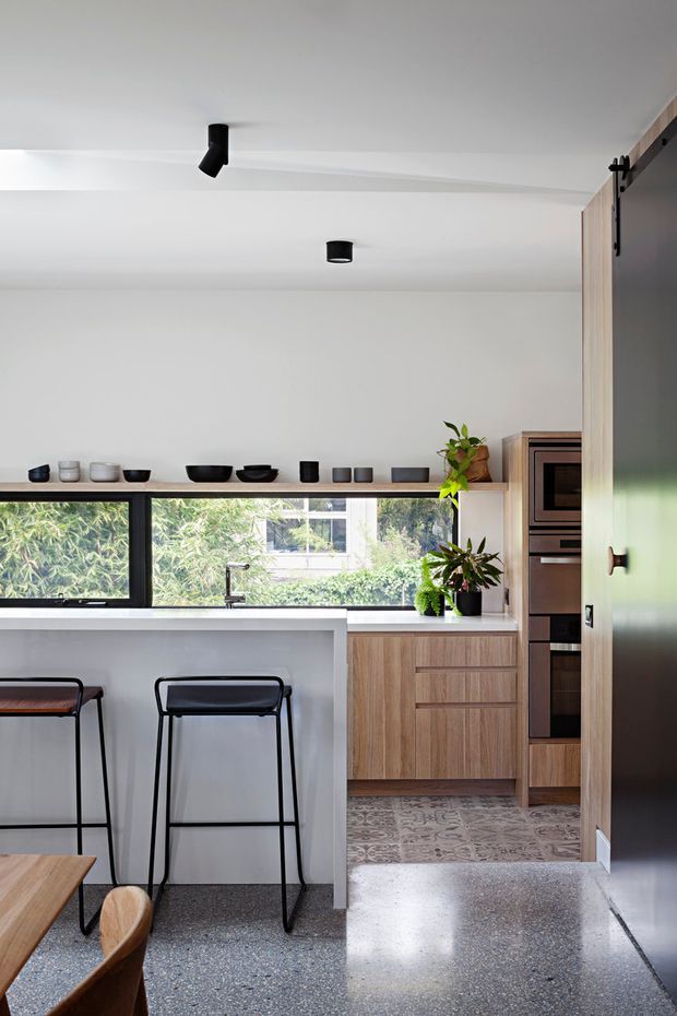A bright, compact contemporary kitchen with a white island and integrated breakfast bar, black-framed horizontal windows, warm wood cabinetry, built-in stainless-steel ovens, and a large dark sliding door. Black stools and track lighting provide contrast, while terrazzo flooring and potted plants add texture and greenery.