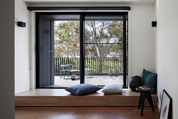 A bright, minimalist interior centered on a large black-framed glazed door opening to a balcony or terrace. A built-in timber window seat with drawers spans the opening, styled with cushions and a small side table and chair. White walls, timber flooring, and strong indoor-outdoor connection create a clean contemporary atmosphere.