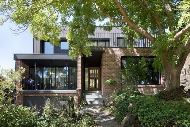 Front exterior view of a contemporary multi-level brick residence, partly screened by mature trees and landscaped garden. The facade combines dark brickwork, extensive black-framed glazing, a projecting flat-roofed entry canopy, and a contrasting upper level clad in vertical light-grey metal panels. A recessed entry is reached by a short stair and walkway.