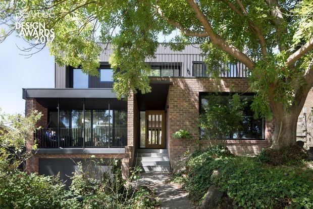Street-facing view of a contemporary multi-level residence integrated into a leafy garden setting. The facade combines exposed dark brick, dark metal cladding and extensive black-framed glazing, with a projecting upper volume and recessed entry reached by a short stair. Mature trees and dense planting partially screen the house.