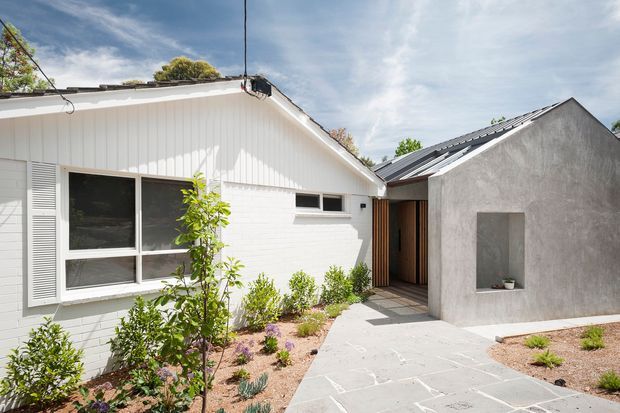 Bright exterior view of a low-rise residential dwelling with a white vertical-panel and painted-brick wing adjoining a rough-rendered grey gabled volume. A sheltered timber-lined entry sits between the two forms, opening onto a pale stone-paved path bordered by low drought-tolerant planting. The composition shows a simple contemporary extension or courtyard-facing facade under pitched metal roofs.