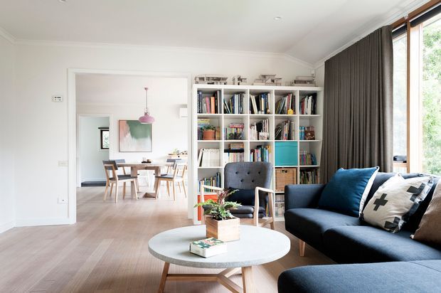 Bright, refined living room with a deep blue sectional sofa, round white coffee table, upholstered occasional chair, full-height dark curtains, and a large built-in white bookshelf filled with books and decor. Pale timber flooring and white walls create an airy setting. A dining area is visible through the wide internal opening, with generous glazing along the right side.